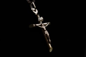 Polished silver crucifix pendant suspended on a chain against a black background, with a detailed corpus figure, INRI tablet at the top of the cross, and a small oval medallion on the left arm, lit with dramatic side lighting