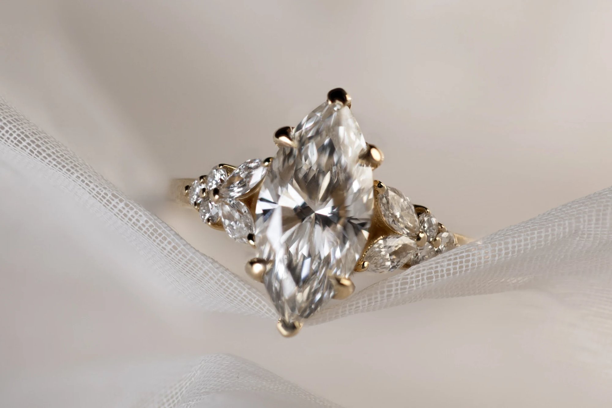 Large marquise-cut diamond ring in yellow gold, viewed from above, resting on white tulle fabric. The center stone is flanked by clusters of marquise and round accent diamonds on either side of the band