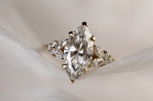 Large marquise-cut diamond ring in yellow gold, viewed from above, resting on white tulle fabric. The center stone is flanked by clusters of marquise and round accent diamonds on either side of the band