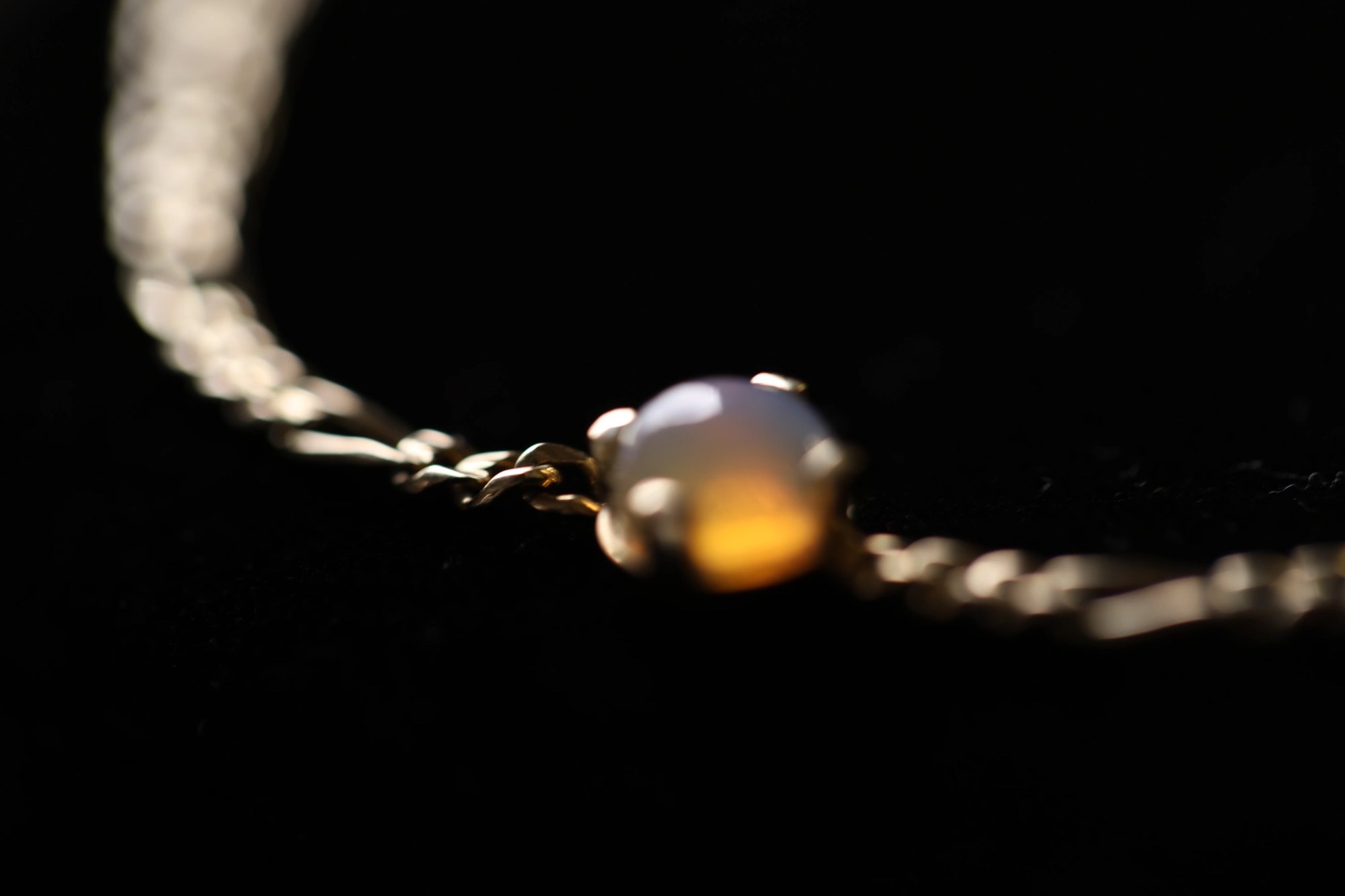 Opal bracelet against a black background with dramatic spotlight lighting, the cabochon opal glowing with warm amber and milky white tones, suspended on a fine gold chain