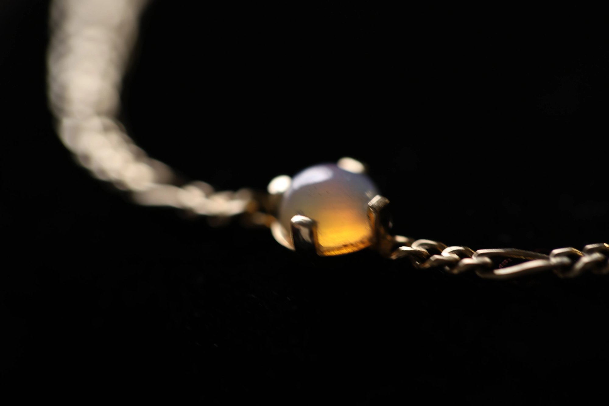 Opal bracelet against a dark background showing the cabochon opal's warm amber glow and white iridescence in a gold prong setting, with the fine chain visible on either side