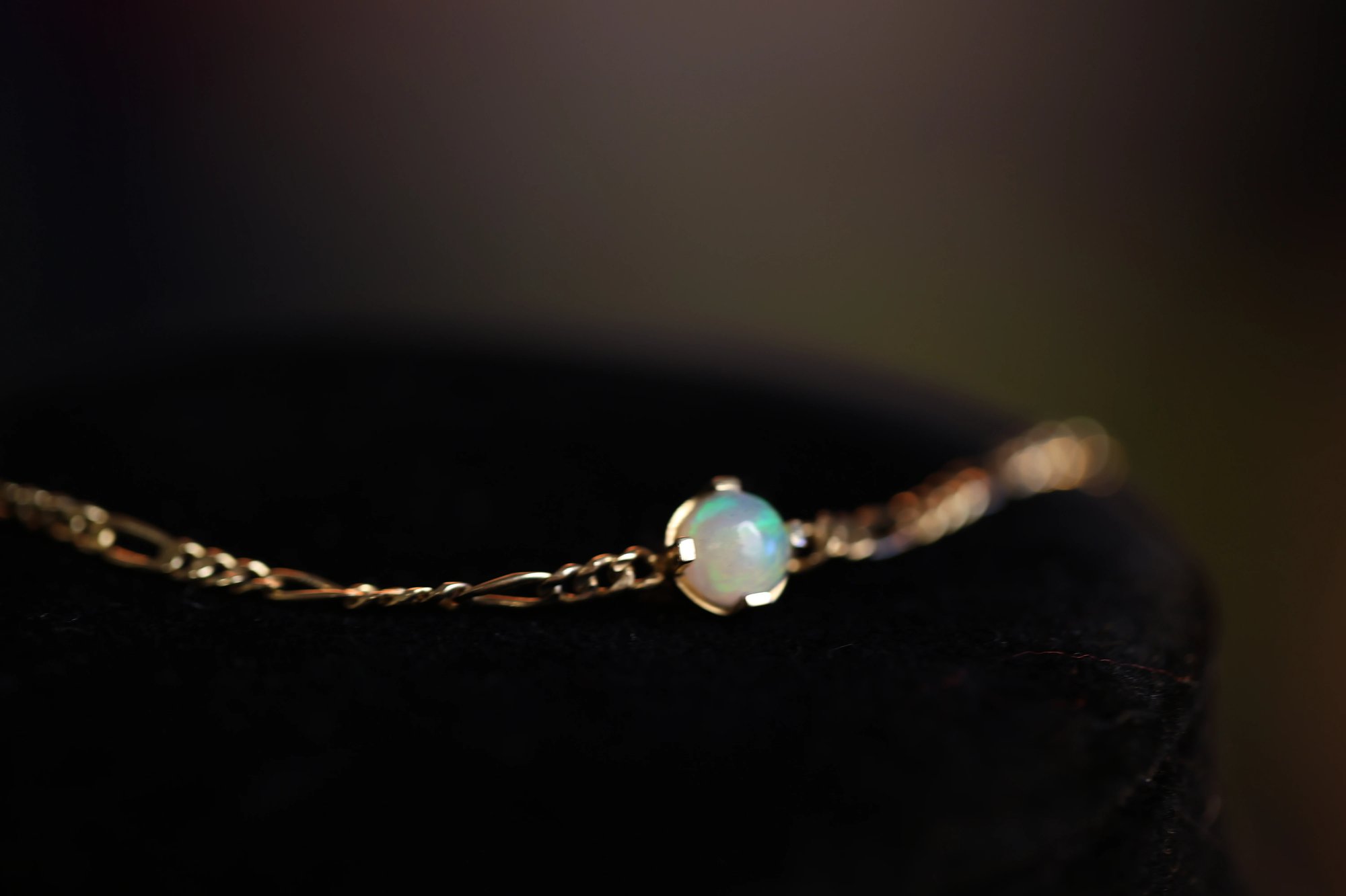 Opal bracelet on dark velvet, showing a round cabochon opal with vivid green and blue play-of-color, set in a four-prong yellow gold setting on a delicate link chain