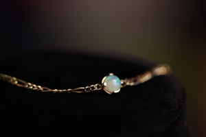 Opal bracelet on dark velvet, showing a round cabochon opal with vivid green and blue play-of-color, set in a four-prong yellow gold setting on a delicate link chain