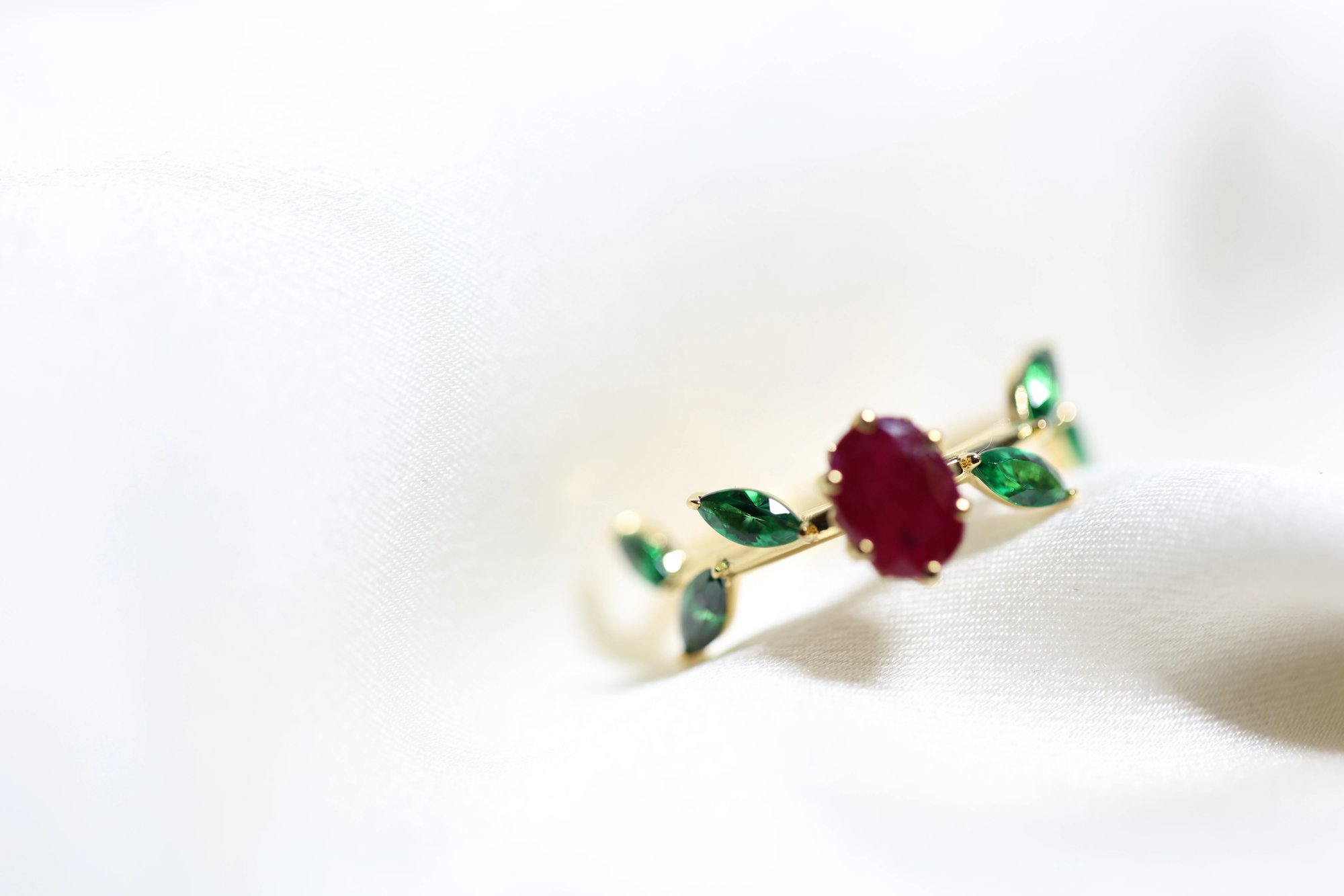 Ruby and emerald ring in yellow gold on white satin, viewed from a slightly different angle showing the ruby center stone and the leaf-shaped emerald accents along the band
