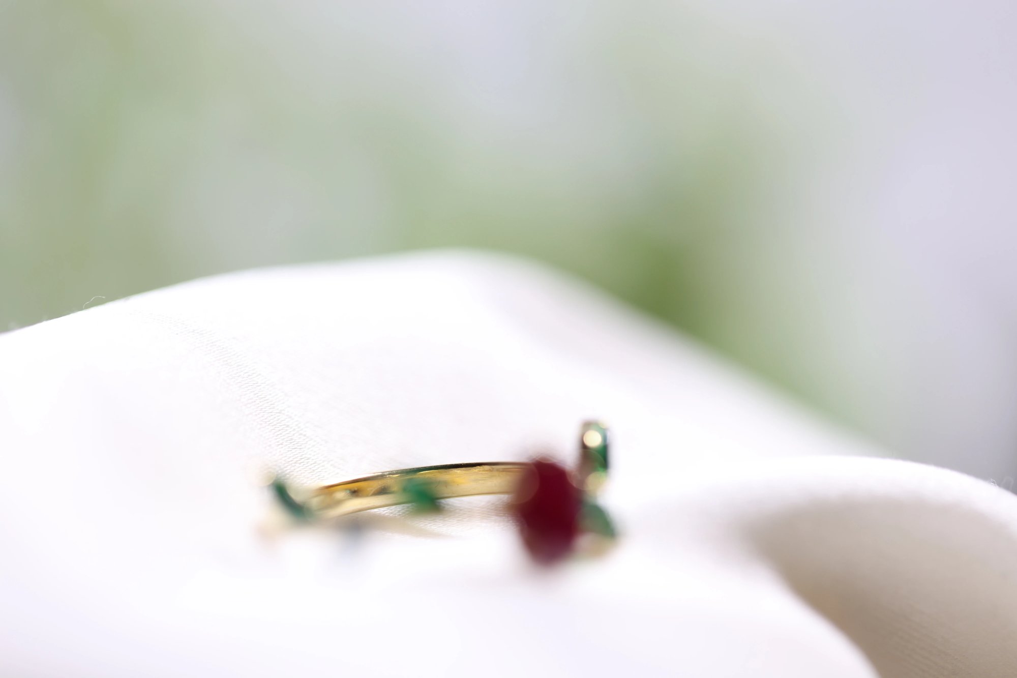 Ruby and emerald ring resting on a white surface, shot as an extreme close-up with shallow depth of field showing the gold band and a hint of the ruby and emerald stones