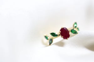 Ruby and emerald ring in yellow gold resting on white satin, showing an oval deep red ruby center stone flanked by marquise-cut green emerald accent stones arranged like leaves along either side of the open band