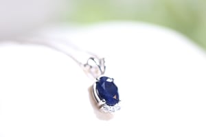 Sapphire pendant close-up showing the oval deep blue sapphire stone, white metal prong setting, and three round diamond accents at the base of the pendant, photographed against a soft white background