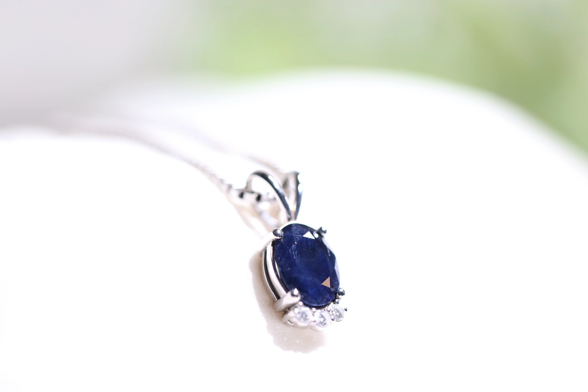 Sapphire pendant with an oval deep blue sapphire in a white metal setting and diamond accents at the base, photographed close-up against a white surface with soft bokeh background