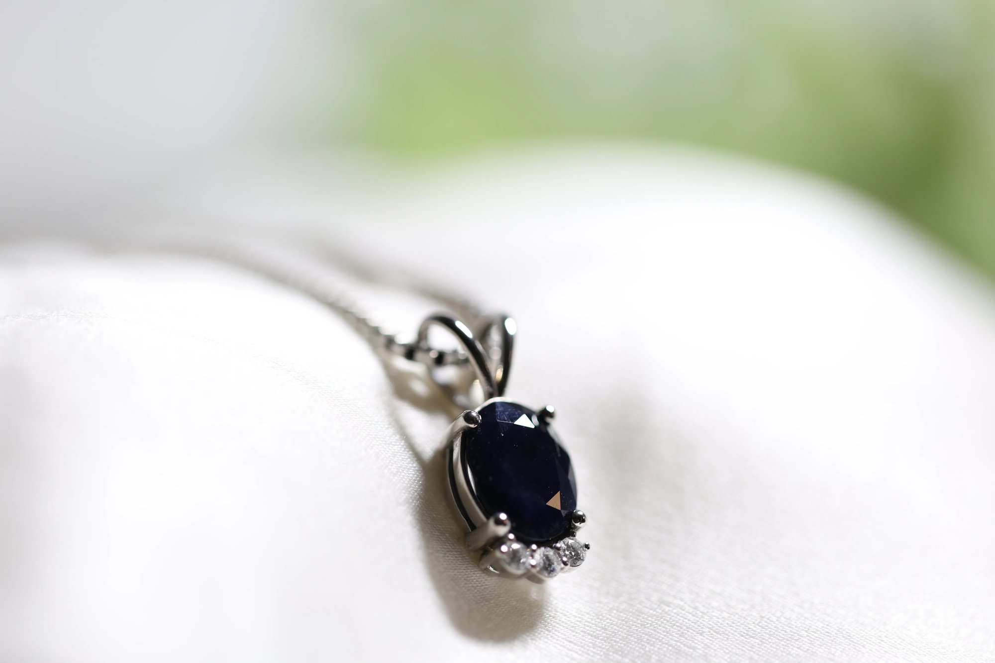 Sapphire pendant on a fine silver chain resting on white fabric, featuring an oval deep blue sapphire in a white metal prong setting with three small round diamond accents below the stone and a decorative scroll bail above