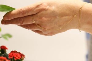 Gold hand chain worn across the back of a hand, showing the delicate fine-link chain running from the wrist to a finger loop with small bezel-set diamond stations at intervals, photographed against a soft white background with red flowers in the foreground