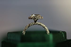 Square cut diamond ring in white metal shown in profile inside a dark green velvet jewelry box, revealing the diamond pavé halo from the side and the arched diamond-set band with its distinctive crossed structure