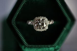 Square cut diamond ring in white metal, photographed from directly above resting in a dark green hexagonal velvet jewelry box, showing a large cushion-cut diamond center stone surrounded by a pavé diamond halo and diamond-set band