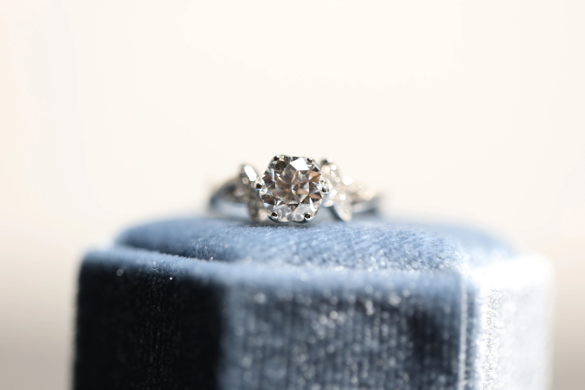 Round brilliant-cut diamond ring in white metal displayed on top of a slate blue velvet ring box, showing the large center stone and flanking accent stones against a neutral background