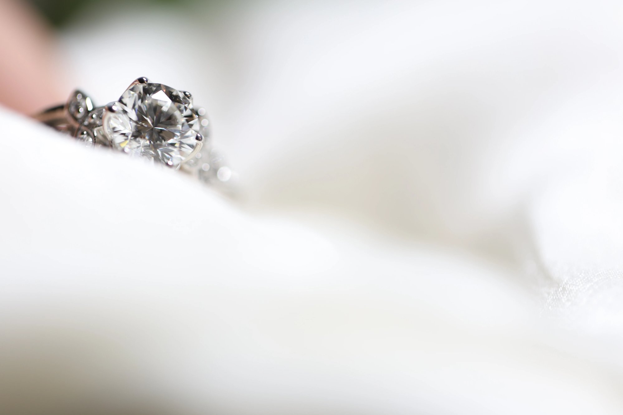 Round brilliant-cut diamond ring in white metal perched on a white surface edge with extreme close-up focus on the large center stone and the decorative scroll detailing of the band shoulders