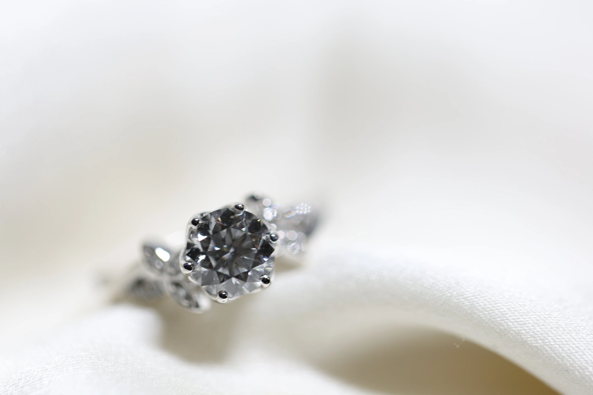 Large round brilliant-cut diamond ring in white metal, photographed from above on white satin, showing the faceted center stone surrounded by marquise and petal-shaped accent diamonds on the band