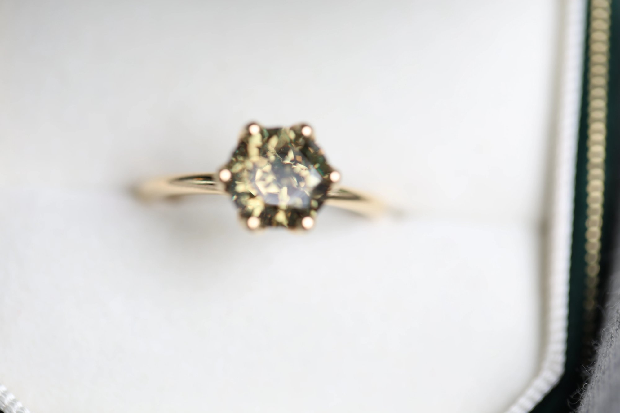 Zircon ring in yellow gold resting in a white-lined dark green jewelry box, featuring a round-cut stone with green-grey coloring in a six-prong hexagonal setting, photographed from slightly above