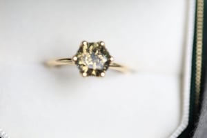Zircon ring in yellow gold resting in a white-lined dark green jewelry box, featuring a round-cut stone with green-grey coloring in a six-prong hexagonal setting, photographed from slightly above