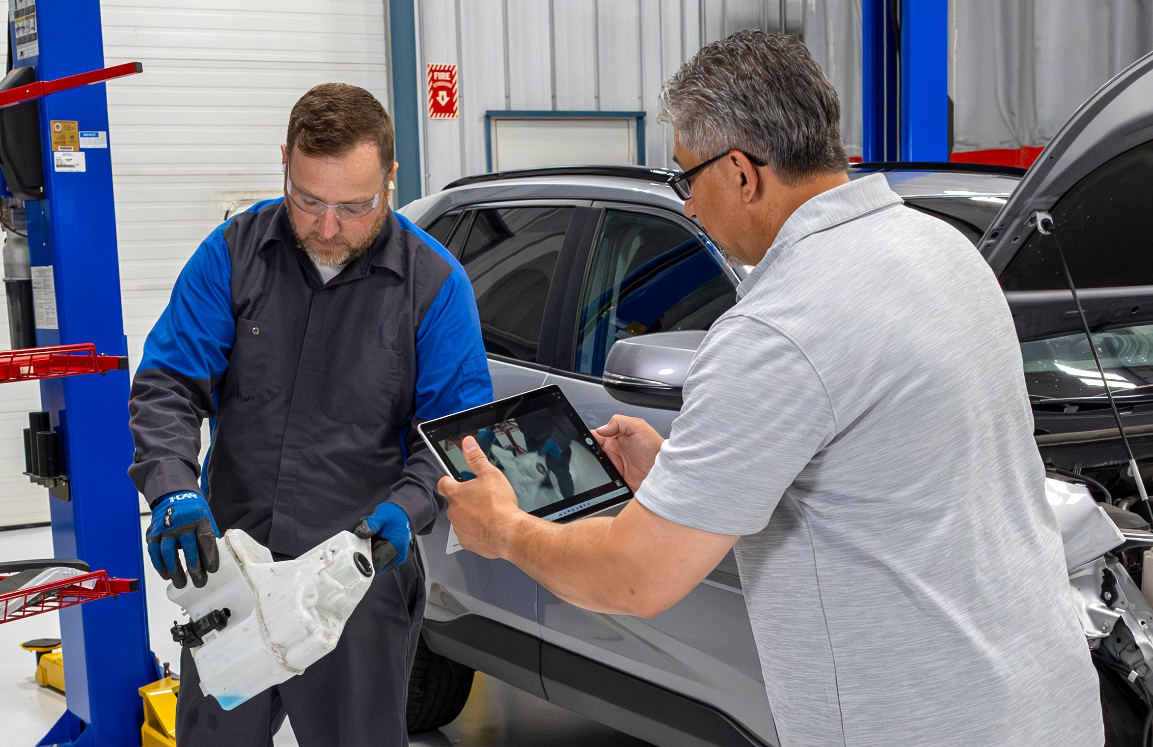 Two collision repair technicians review a damaged wiper fluid reservoir on a tablet while a silver SUV with its hood open sits on a lift in the shop background.