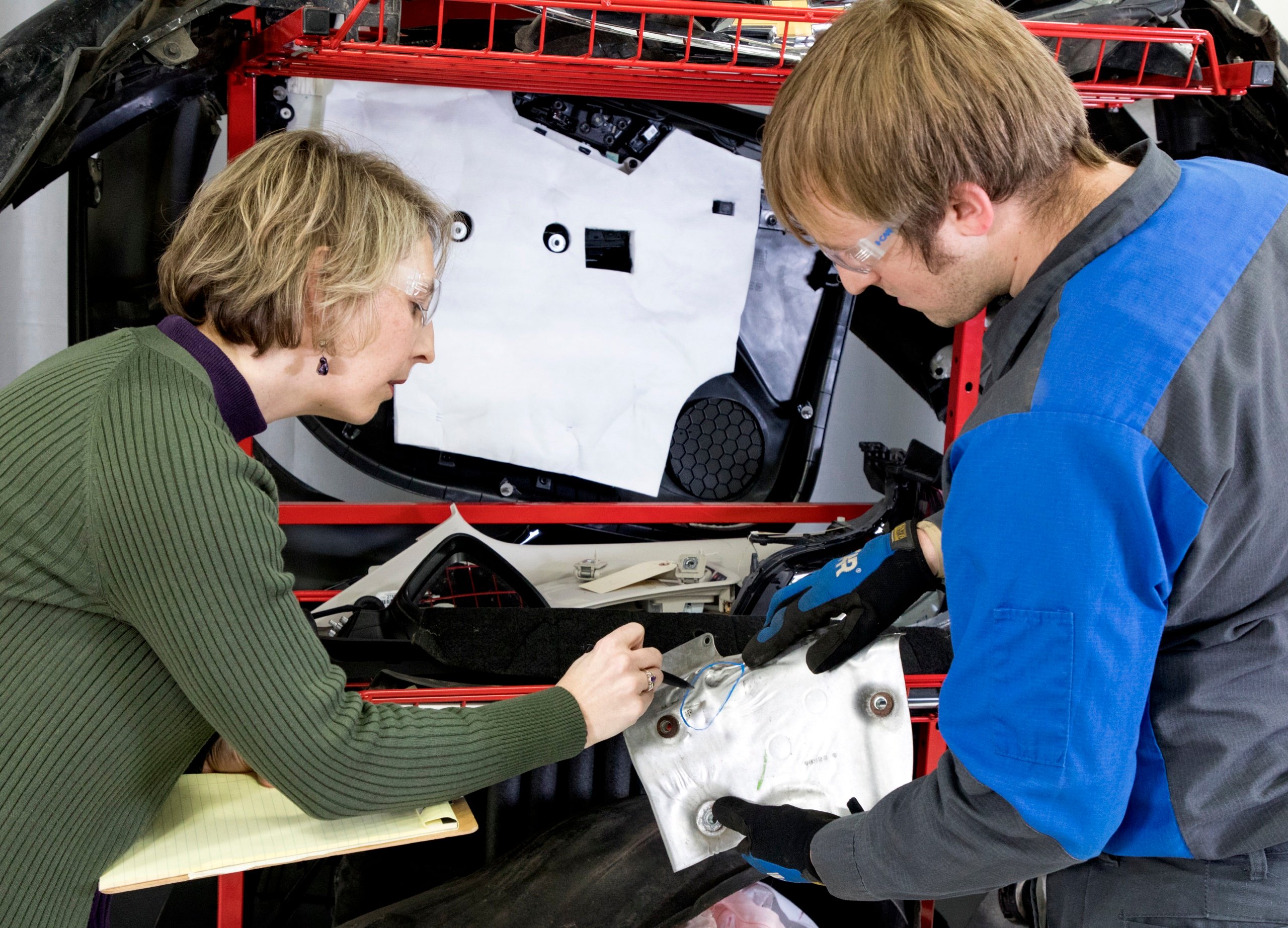 An estimator and a technician examine disassembled front-end parts — a side mirror, headlight, and interior door panel — laid out on a repair rack during a collision damage assessment.