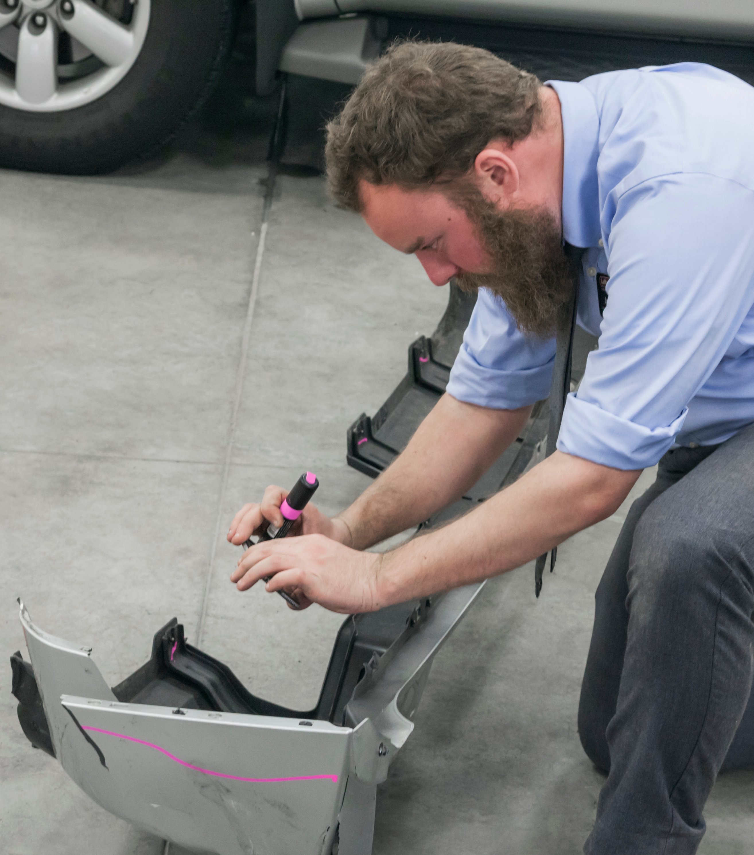 A technician marks repair reference lines on a vehicle's front bumper cover and fascia with a pink paint marker during a collision repair inspection.