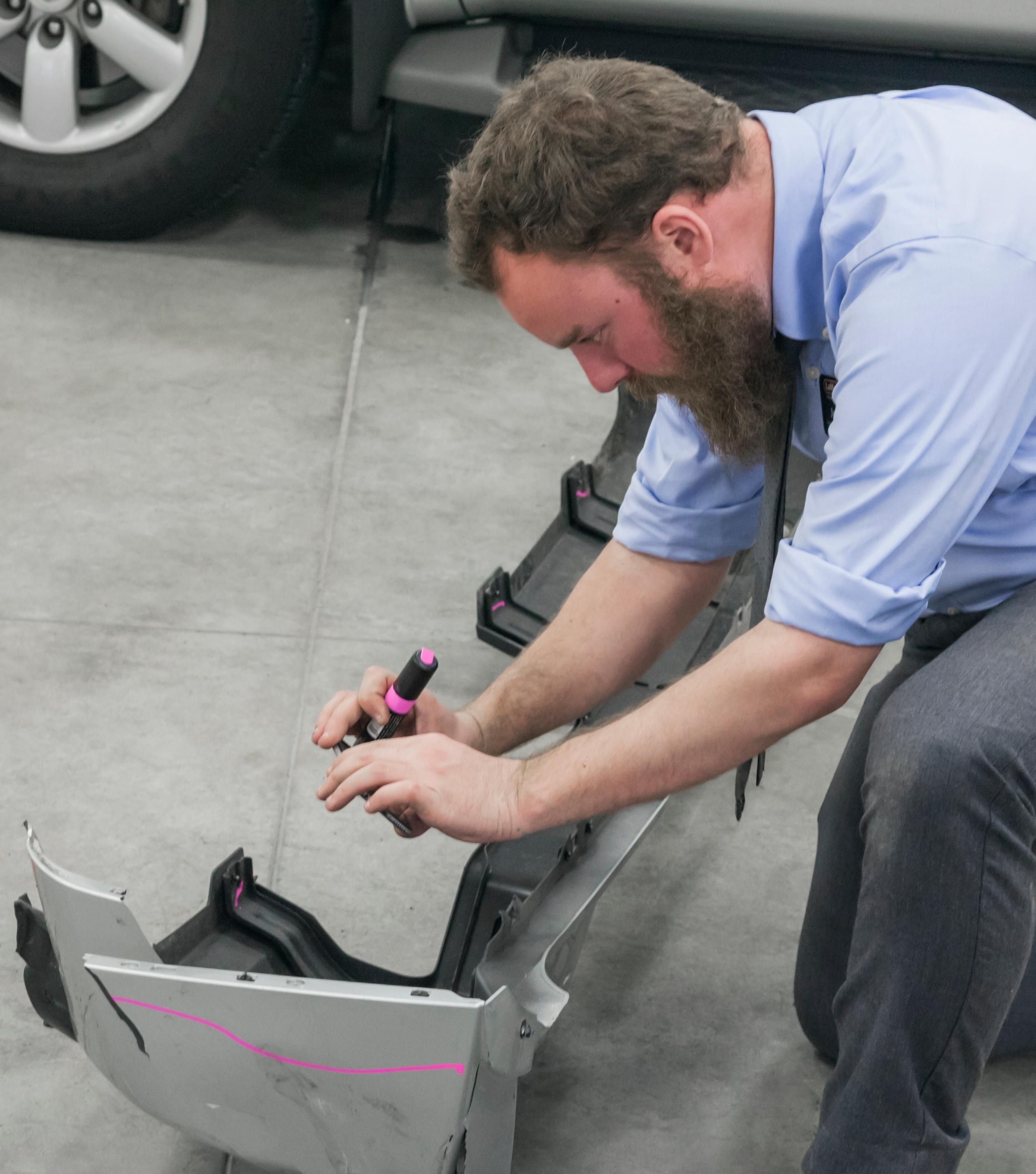 Ein Techniker markiert bei einer Schadenaufnahme mit einem rosafarbenen Lackstift Referenzlinien für die Reparatur auf Stossfängerverkleidung und Frontschürze eines Fahrzeugs.