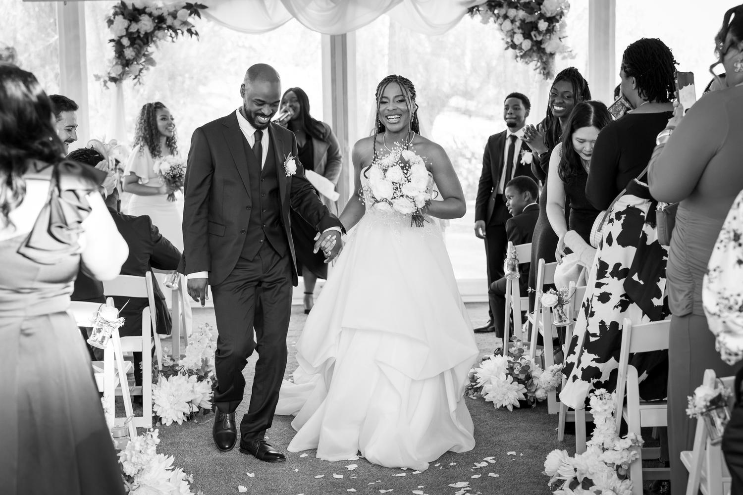 Black and white photo of Chiedza and Foster smiling as they walk back down the aisle