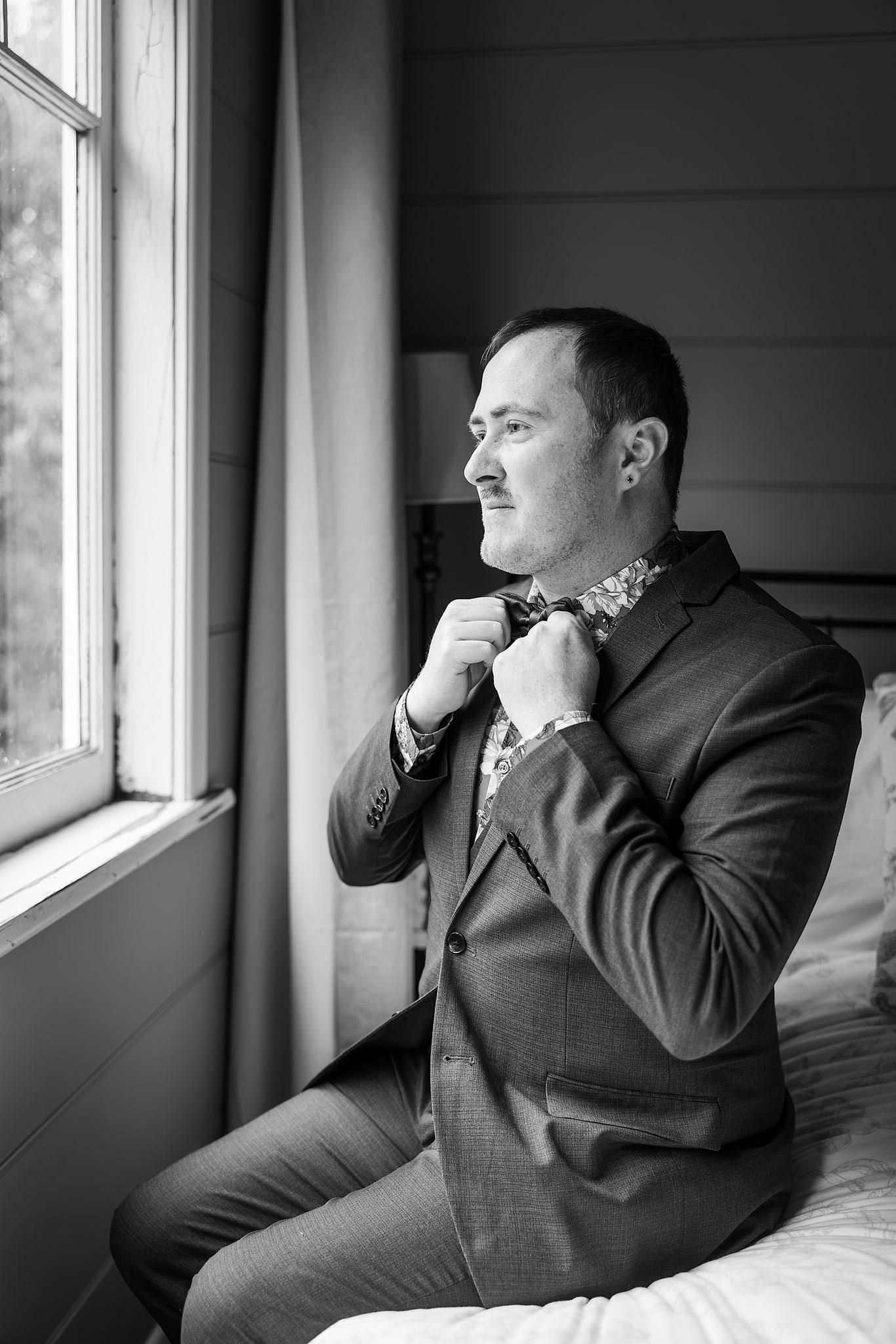 Paul in a suit adjusts his tie in front of a window