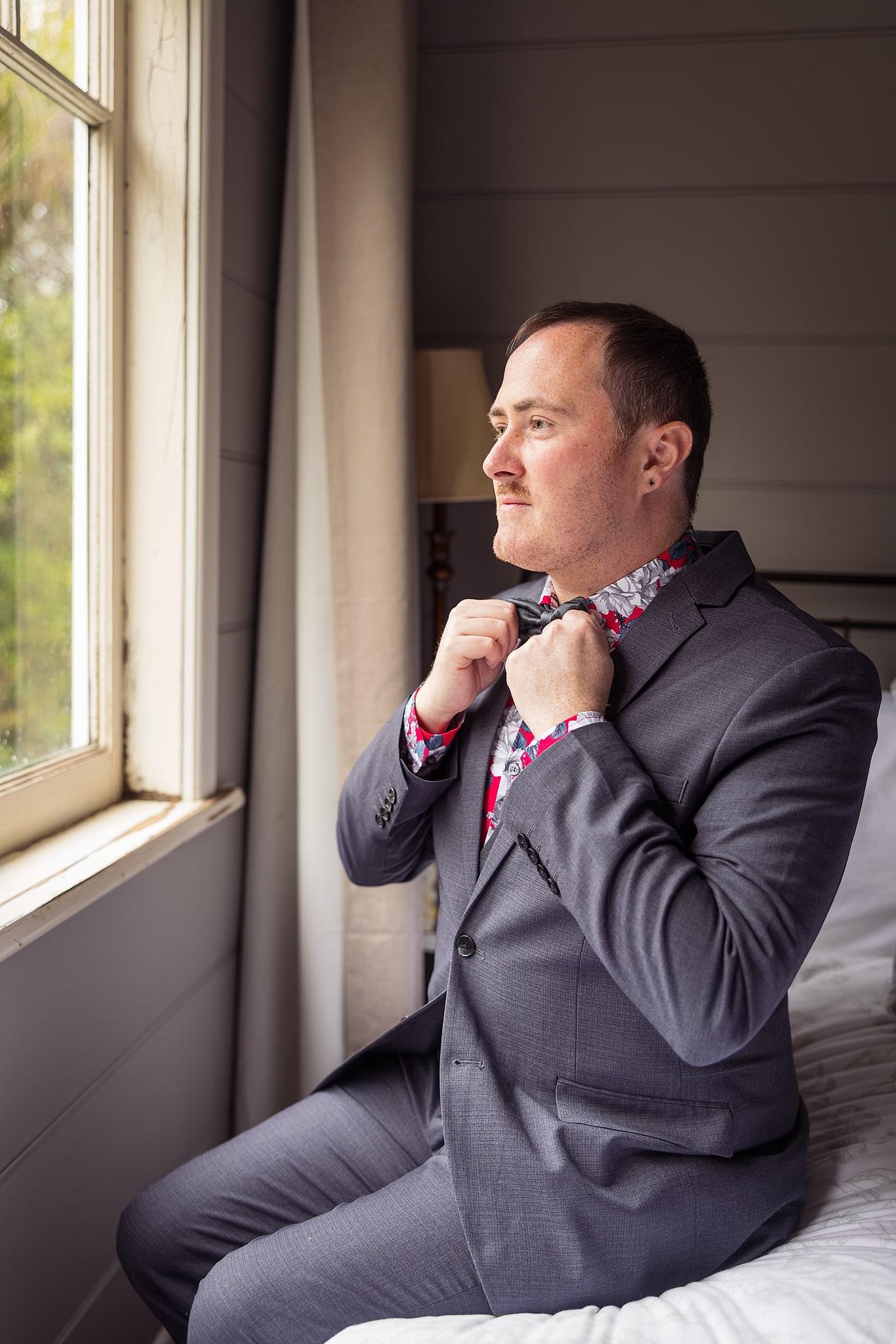 A man in a suit adjusts his tie in front of a window