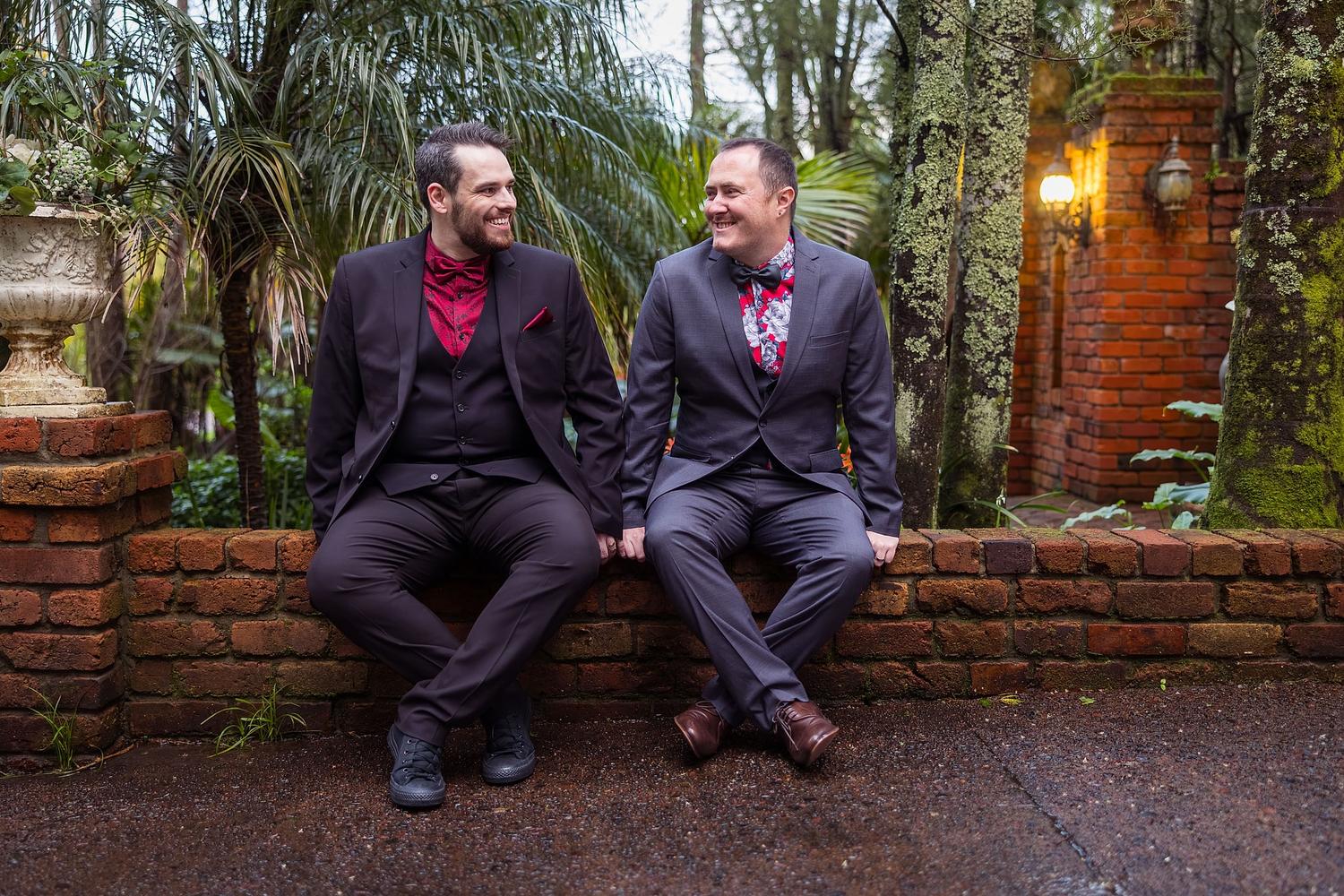Two men in suits sit on a brick wall, smiling and enjoying the moment