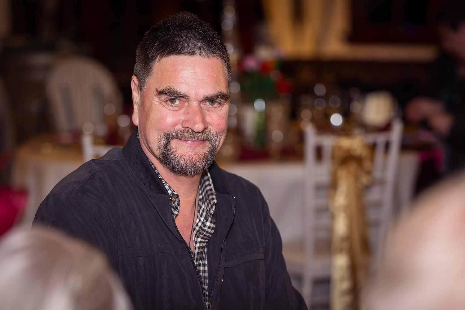 A man in a black jacket is sitting at a table with a wine glass in front of him