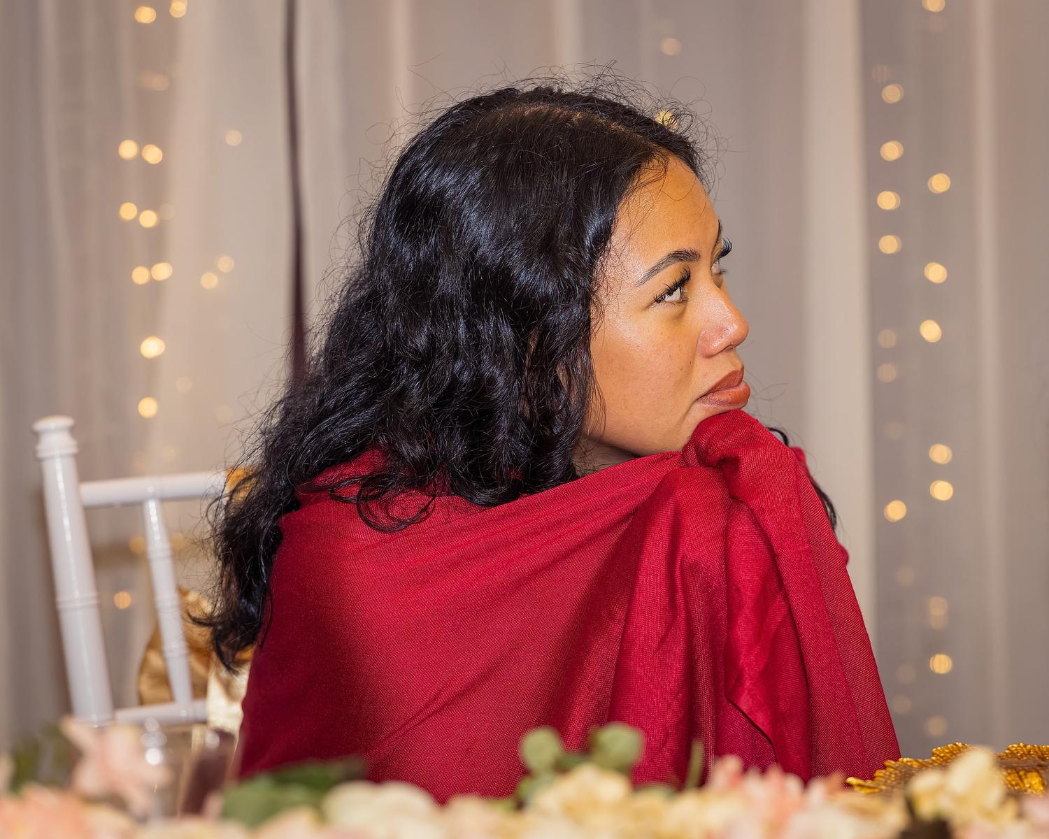 A woman in a red shawl sits at a table with flowers in front of her