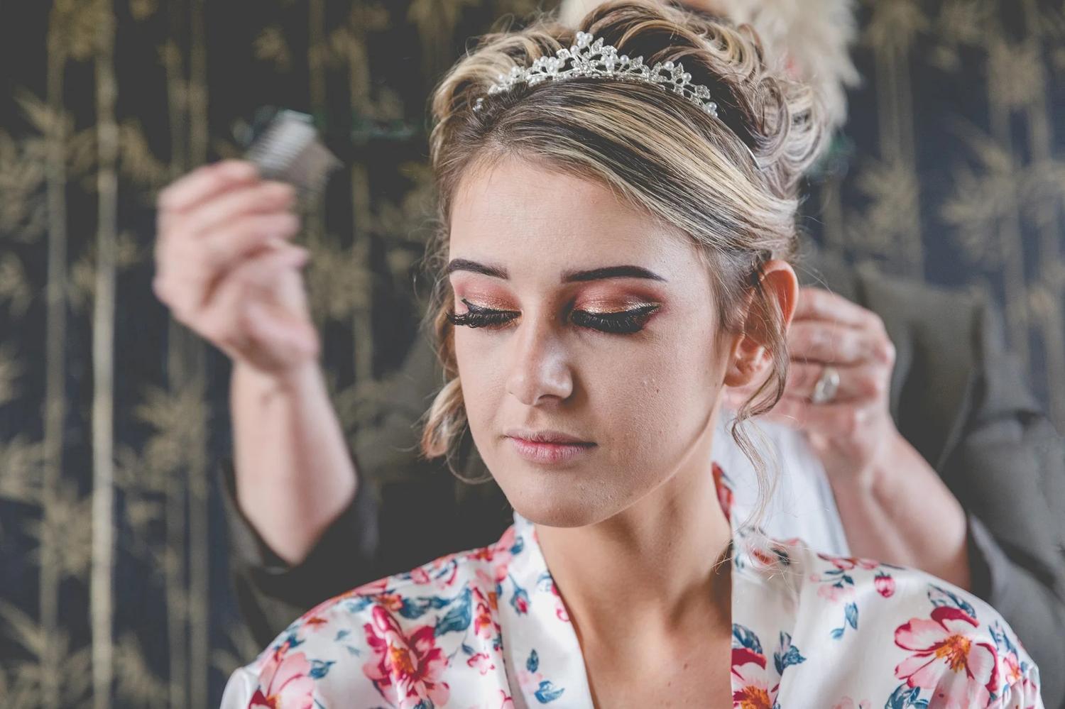 Bride in a floral robe with her eyes closed as her makeup is finished