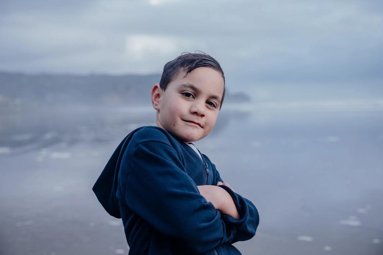 A boy in a hoodie poses with folded arms by the sea
