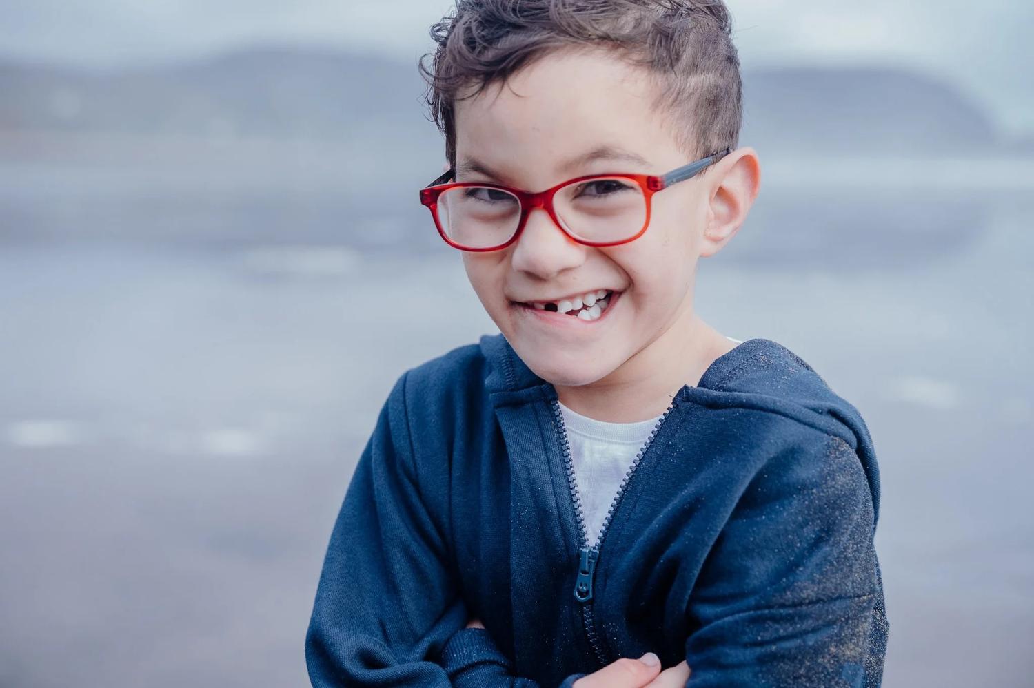 A boy in red glasses grins at the camera on the beach