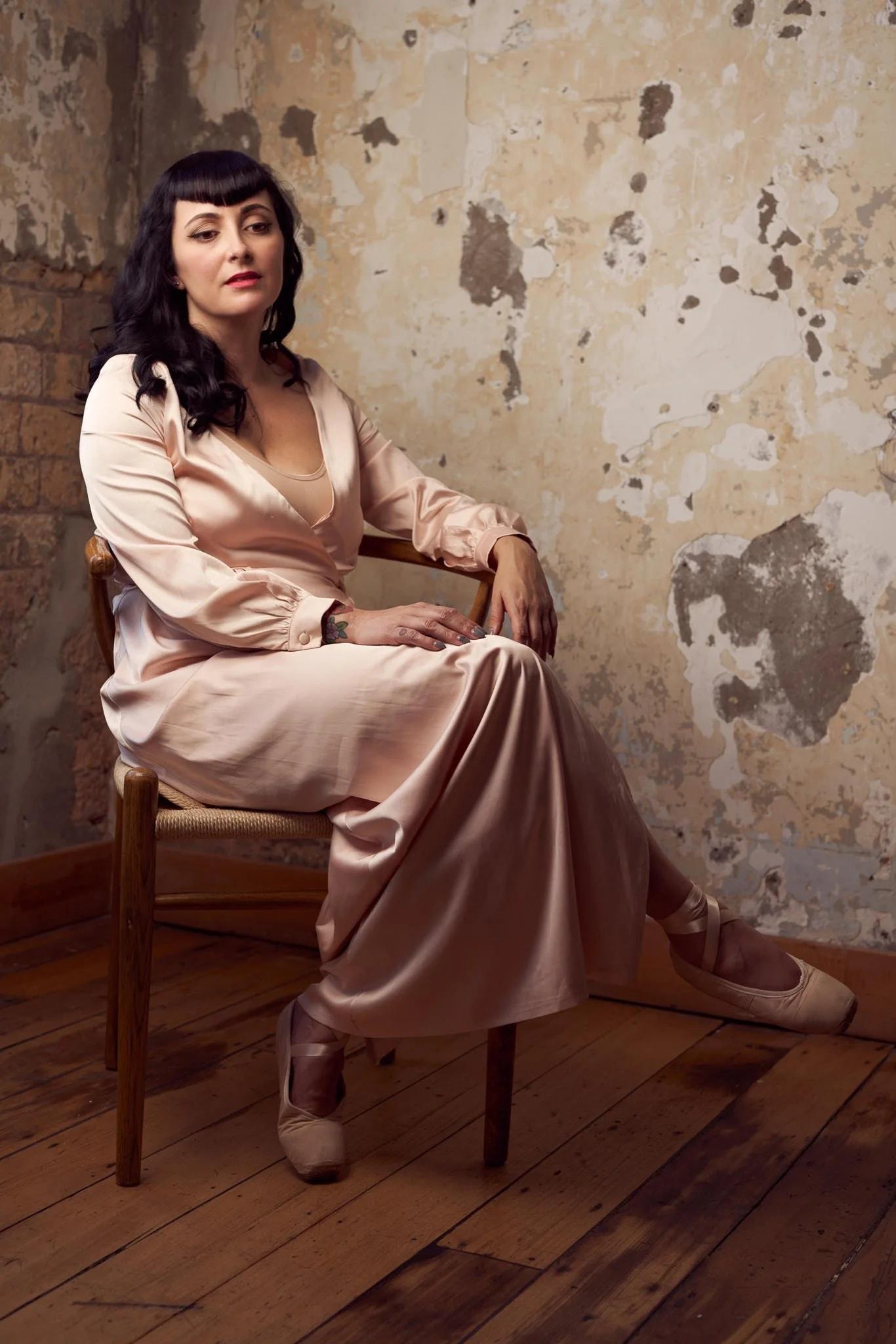 Viv seated on a wooden chair in blush satin and ballet shoes