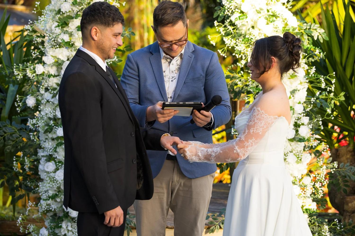 Groom placing the ring on the bride’s finger as the celebrant reads under a floral arch