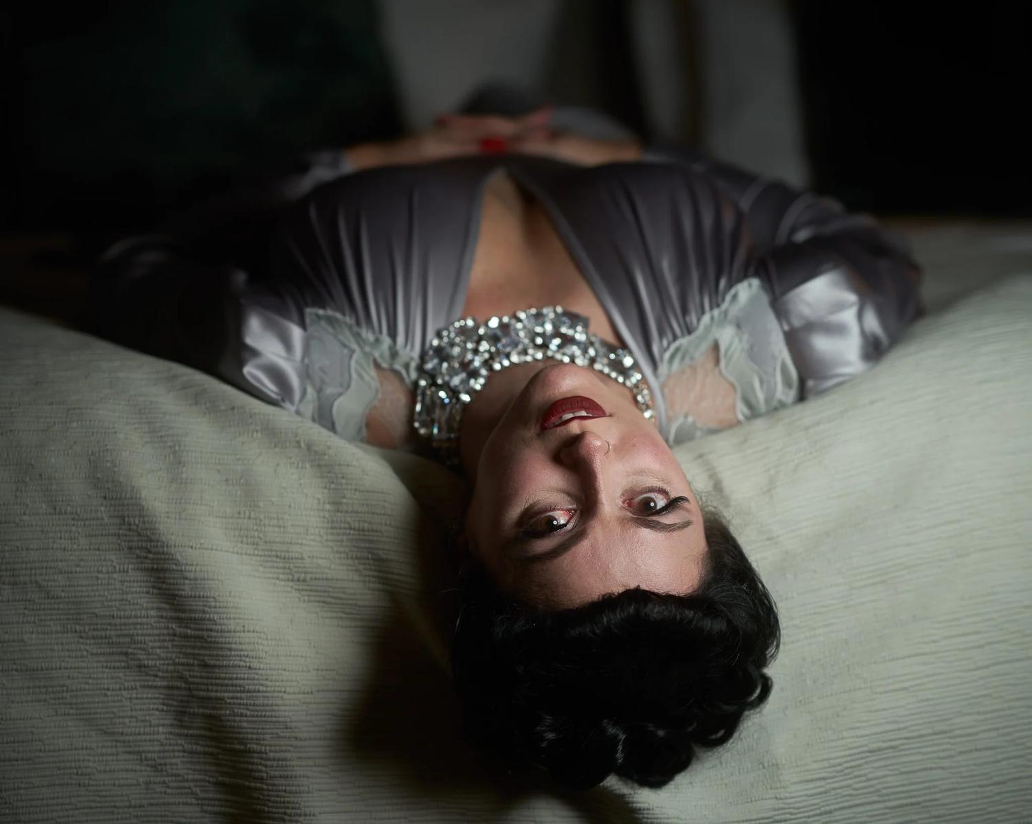 Viv lies upside down on a bed in a silver satin gown