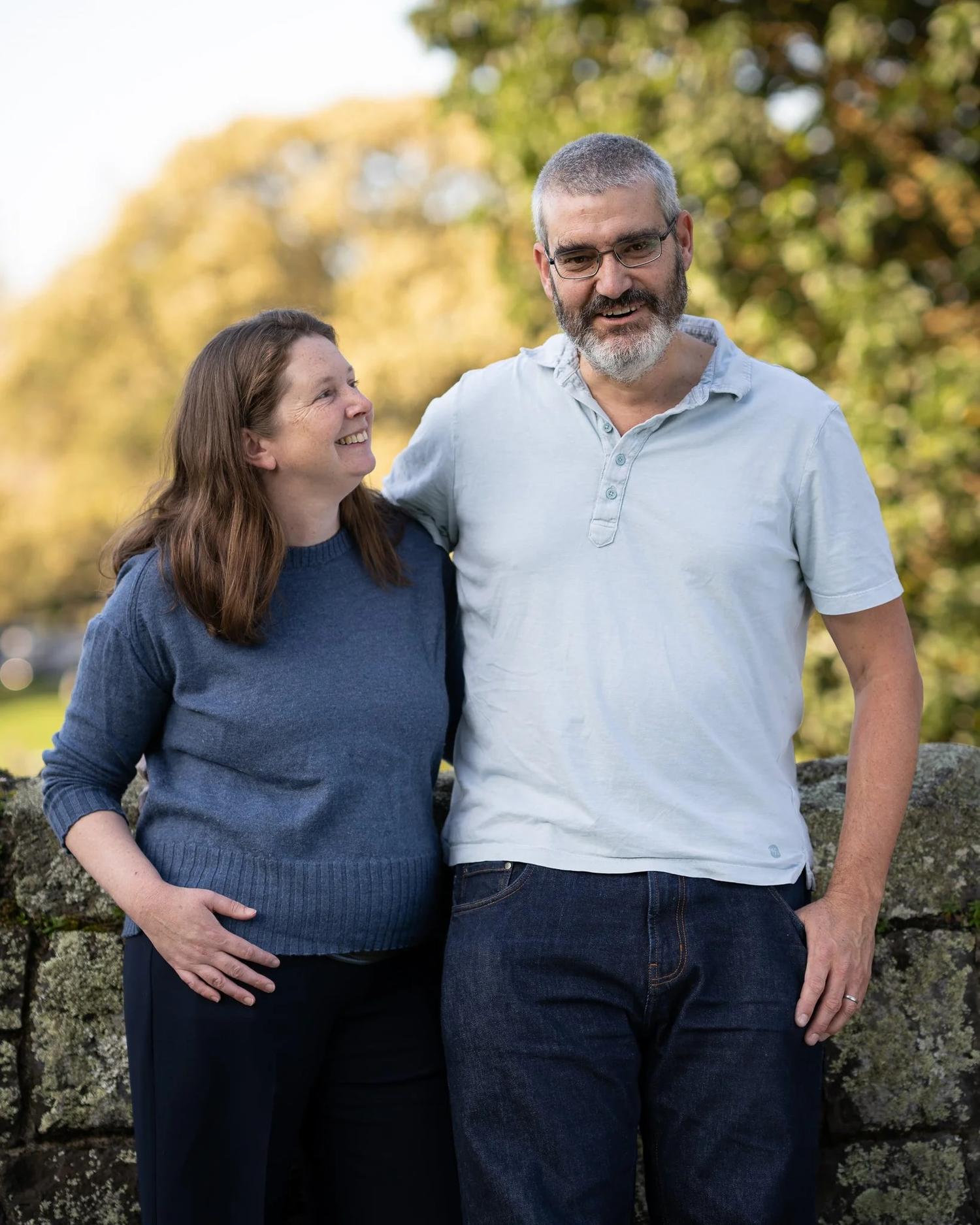 The parents share a smile in front of a stone wall