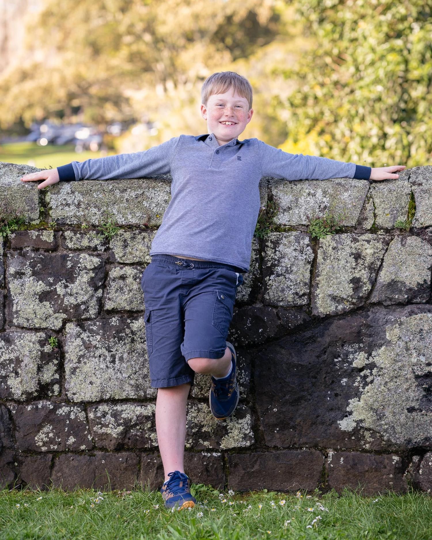 A boy stands on one leg against a stone wall with arms outstretched