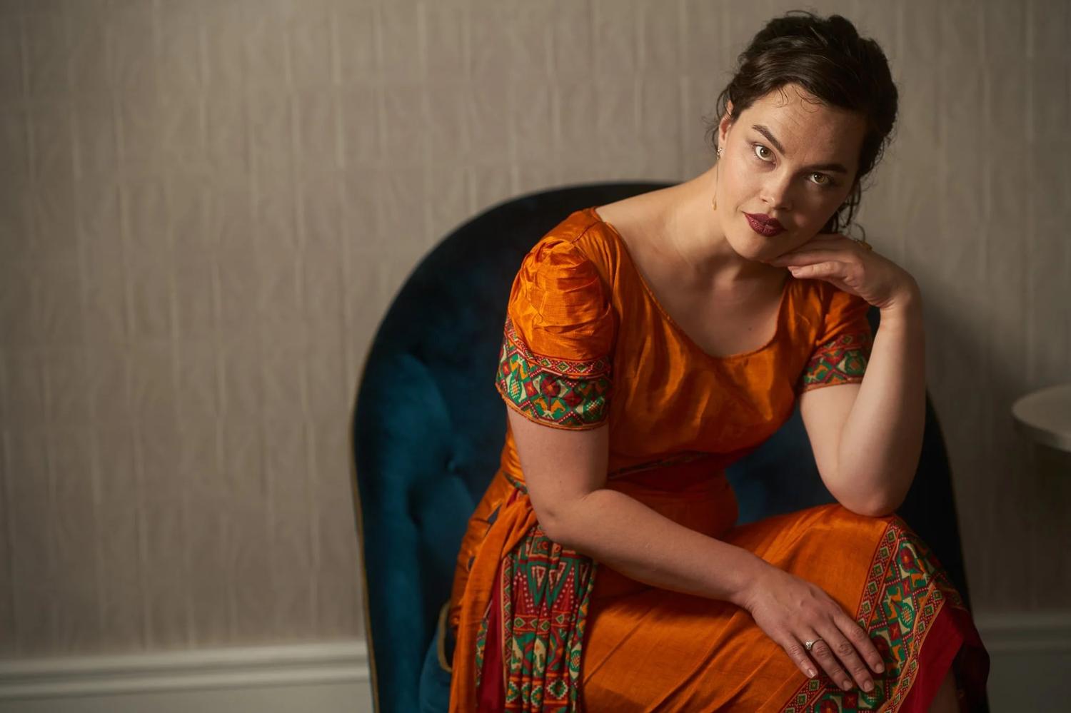 Anita in an orange embroidered dress on a blue velvet chair