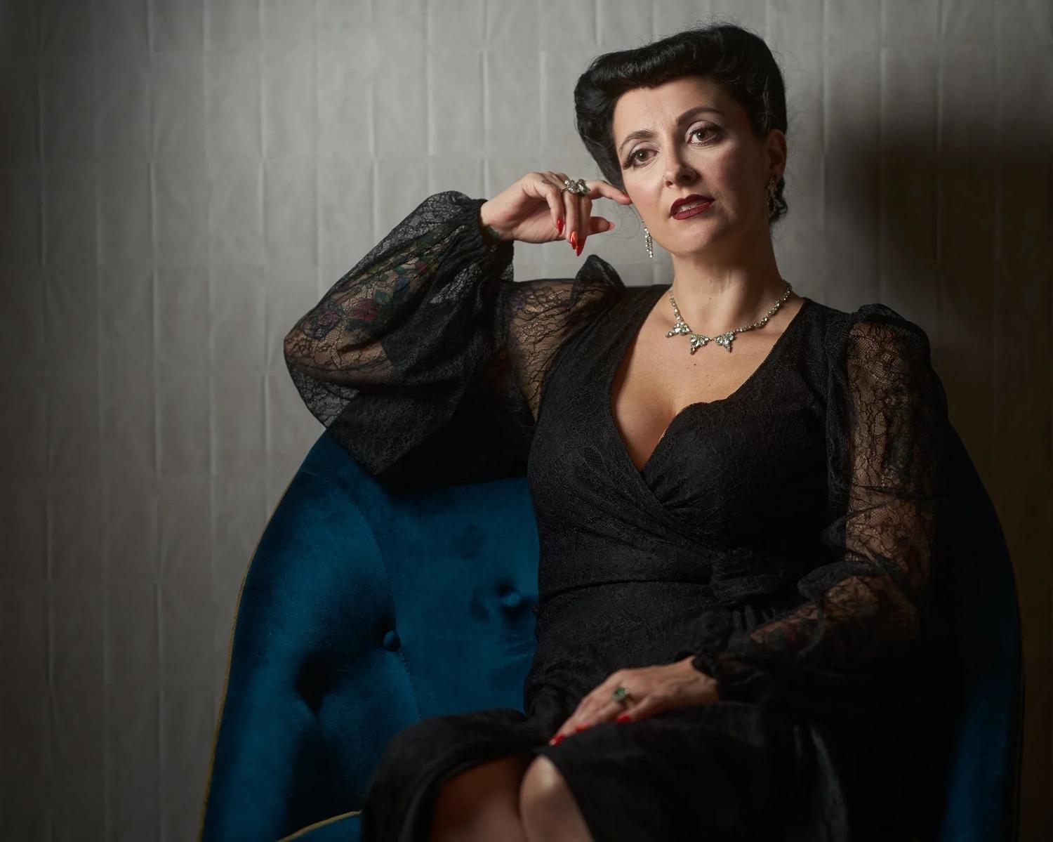 Viv in black lace seated in a teal velvet chair
