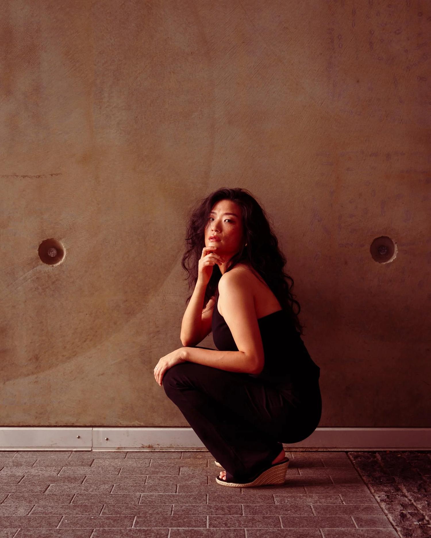 Anna crouches against a concrete wall with her chin on her hand