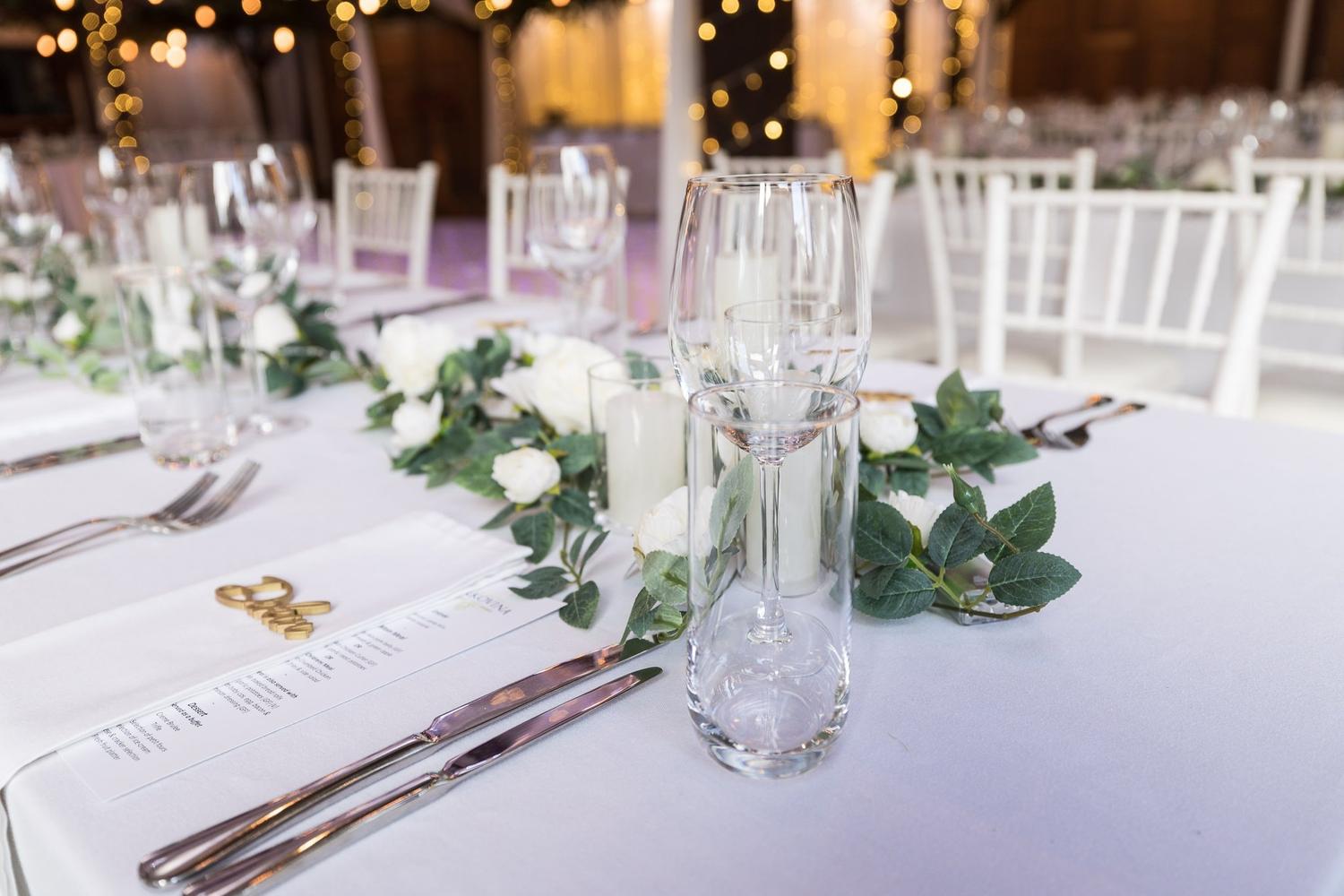 Wedding reception table with white roses, greenery, glasses and a gold table number