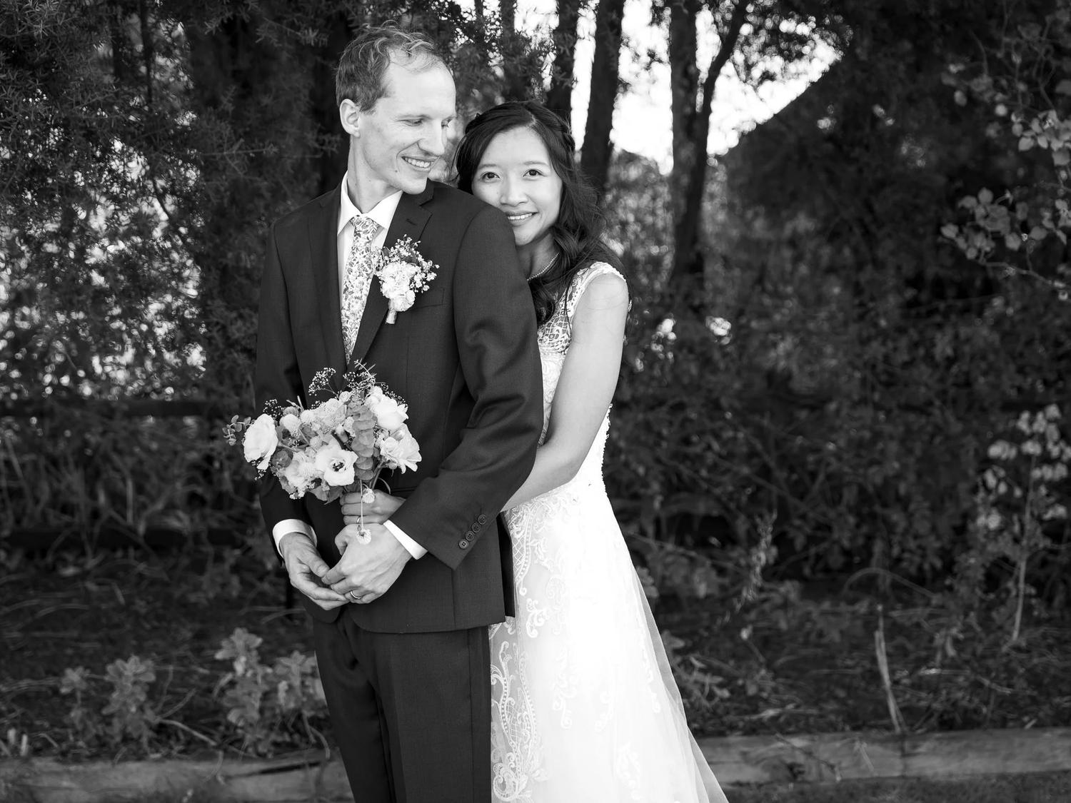 Black and white photo of a bride hugging her groom from behind in a garden