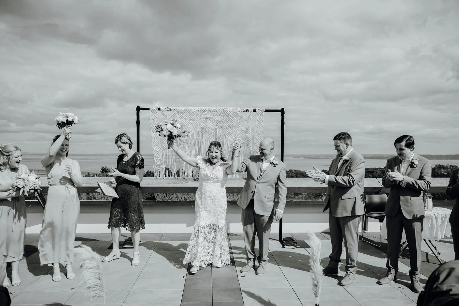 Black and white photo of a couple celebrating with their wedding party on a terrace by the sea
