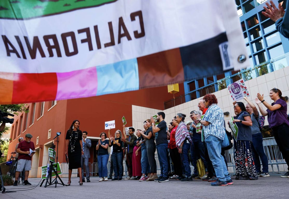 An image of a rally of unions and faculty associations, with people carrying placards with slogans. A prominently-positioned flag of California is in the front of the image.