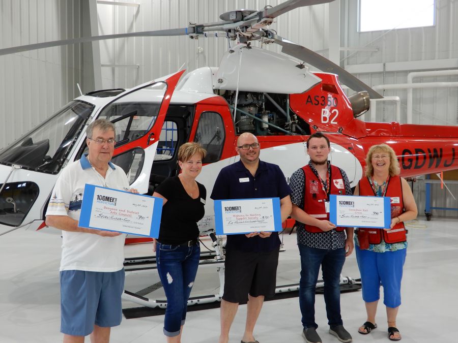 <who>Photo Credit: 100 Men Who Care Penticton</who> Donation recipients pose with their oversized cheques at Topflight Helicopters in May 2019