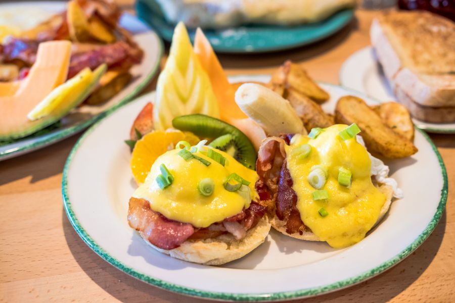 Tomatoes, bacon and green onions Poached eggs on an English muffin covered with hollandaise sauce served with potatoes and fresh fruit.