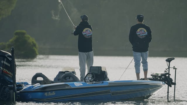 Two anglers in Flatty Classic hoodies casting from the bow, backlit against the treeline