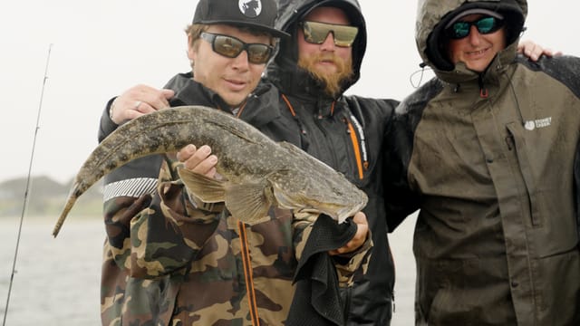 Three anglers in the drizzle, one holding up a solid dusky flathead